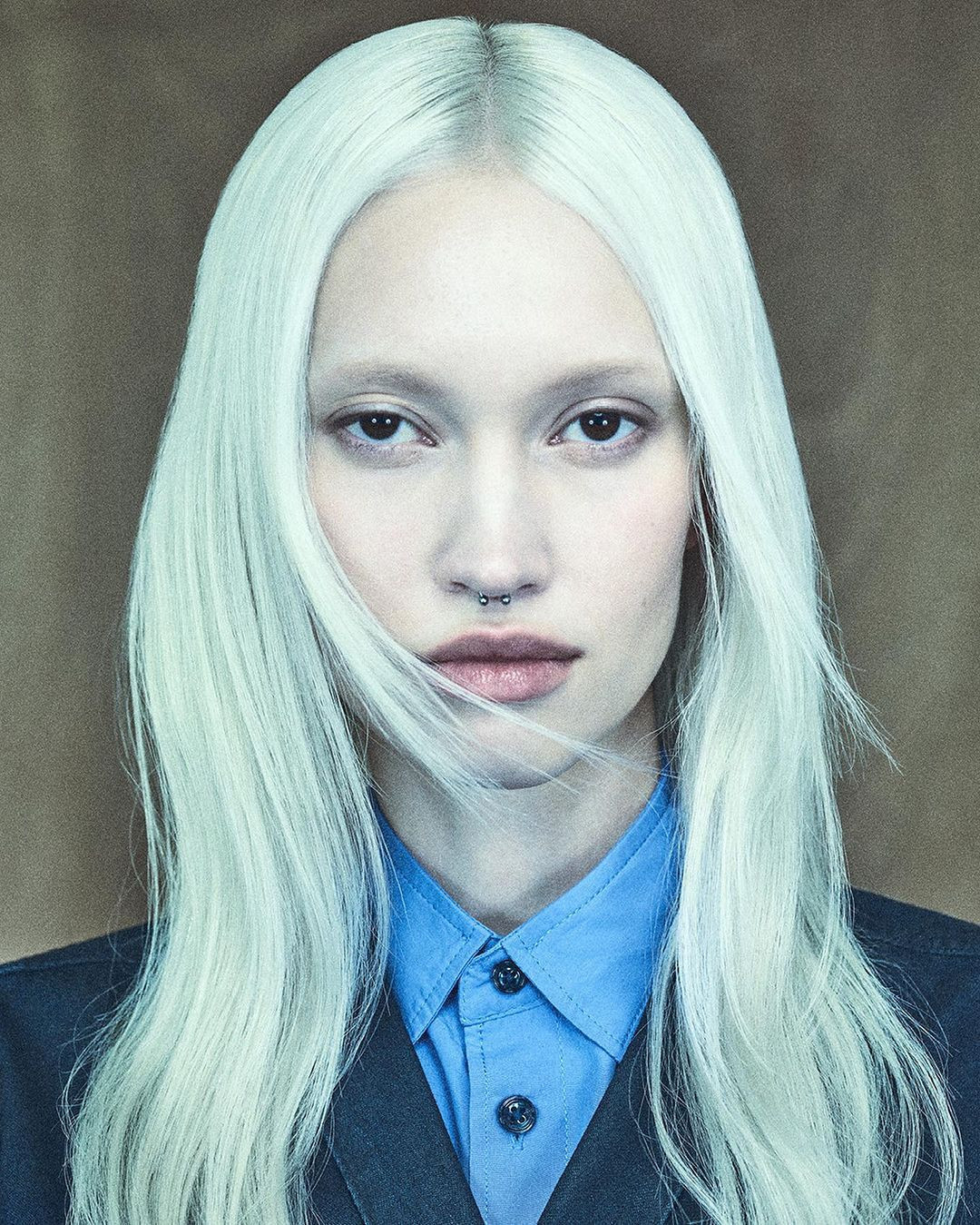 Close-up portrait of an adult female with pale skin, platinum blonde hair, and a blue collared shirt, set against a neutral beige background.