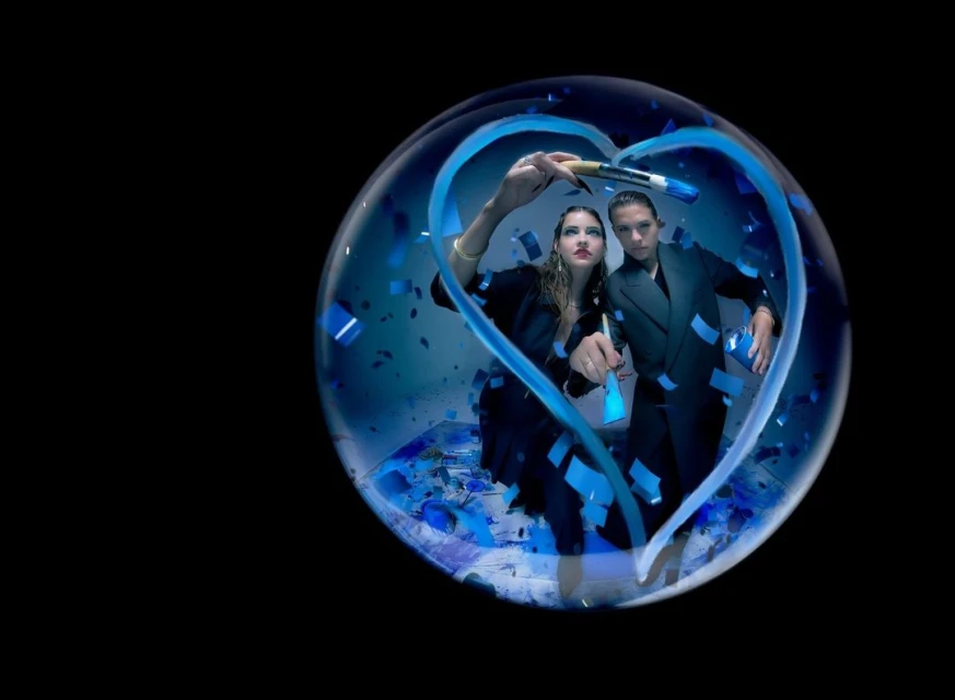 Two adult models in suits pose inside a bubble with blue confetti and paint strokes forming a heart shape against a black background.