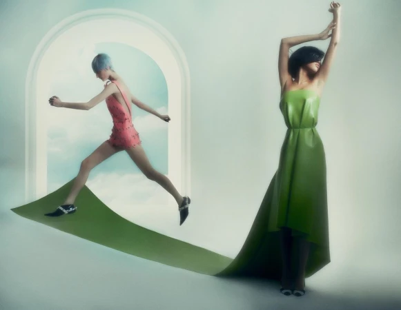 Two women in bright surreal fashion, one leaping through a white arch, the other standing with arms raised, both on a green runway.