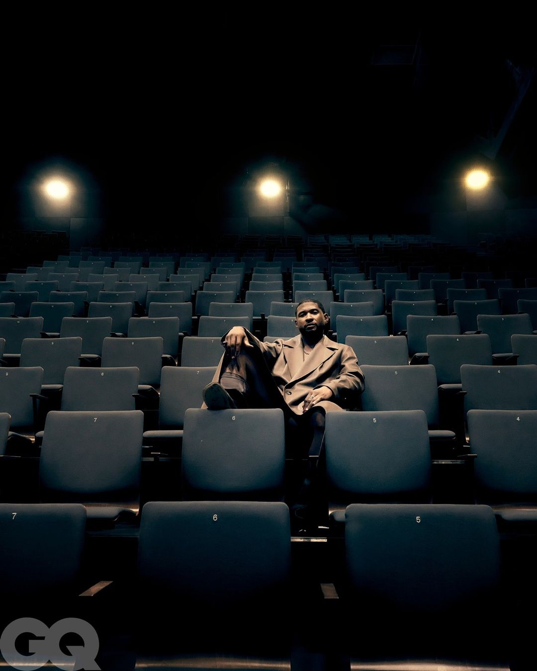An adult male is sitting in a theater with rows of empty blue chairs, wearing a brown jacket, with spotlighting and low angle shot.