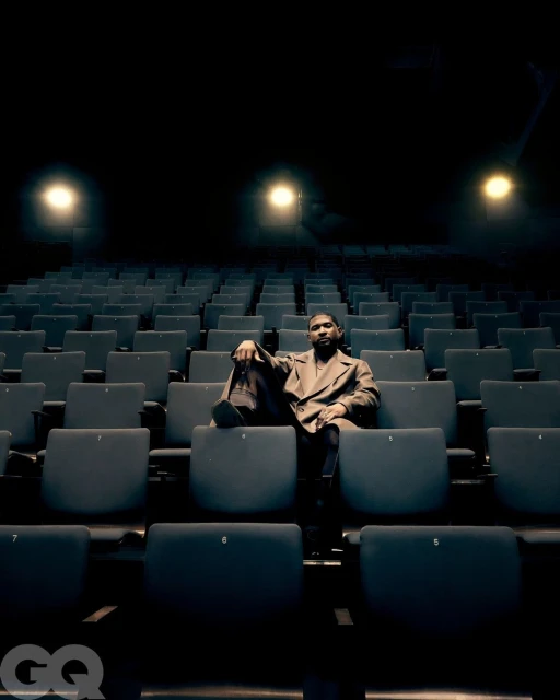 An adult male is sitting in a theater with rows of empty blue chairs, wearing a brown jacket, with spotlighting and low angle shot.