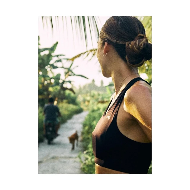 Woman in athletic wear stands on a path in a tropical outdoor location, with a dog and scooter in the background.