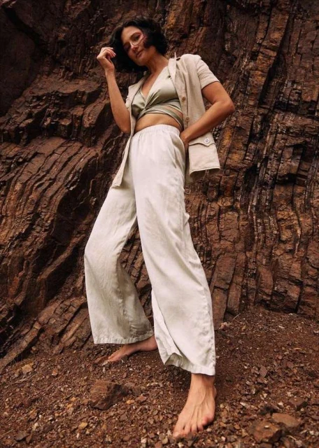 A woman in casual, light-colored clothing stands barefoot on rocky ground against a textured rock formation in an outdoor setting.