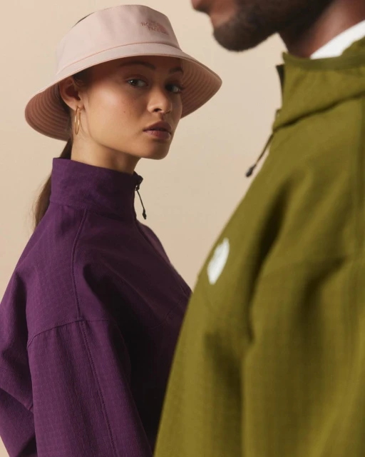 Two people wearing sporty jackets stand indoors against a plain background. One wears a sun visor, and the other wears a green jacket.