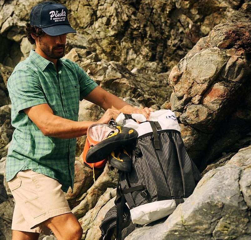 A man in a cap and green shirt puts a climbing shoe in a backpack, outdoors among rocks and boulders on a sunny day.