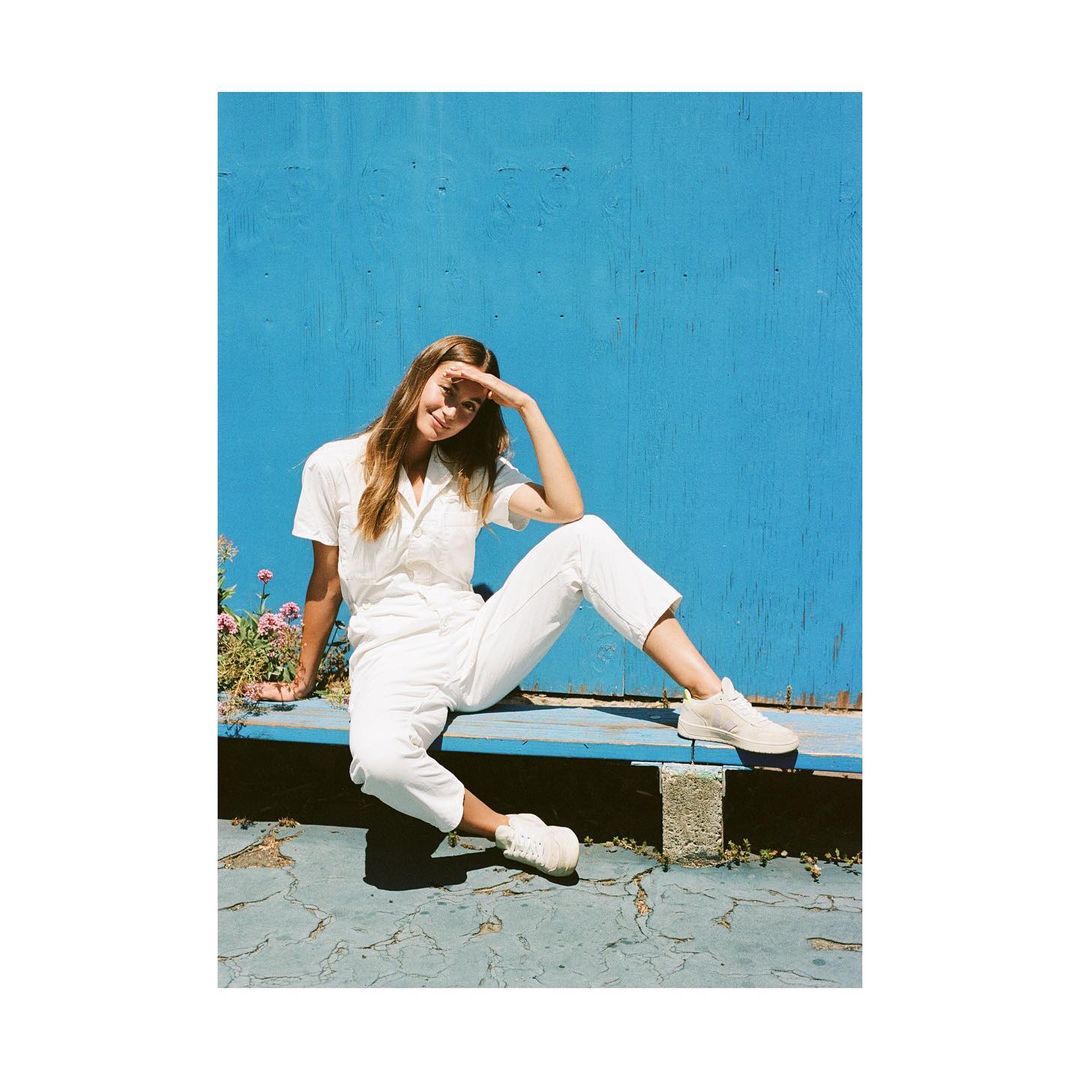 Young woman in white shirt and pants poses against a blue wall outdoors, sitting on a low bench with flowers nearby in natural light.