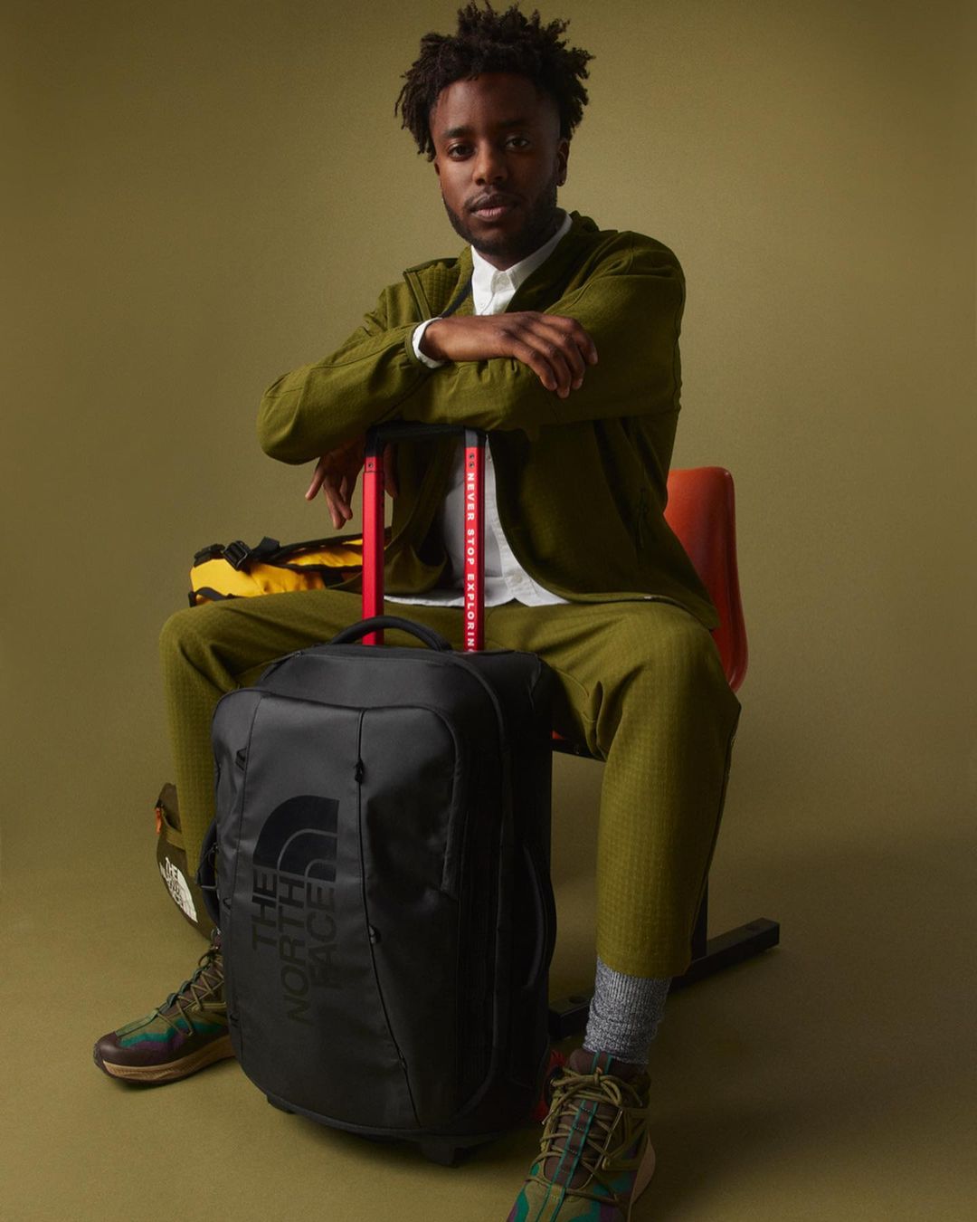 A seated adult Black man in a green suit with a black travel bag and The North Face branded gear, against a neutral green backdrop.
