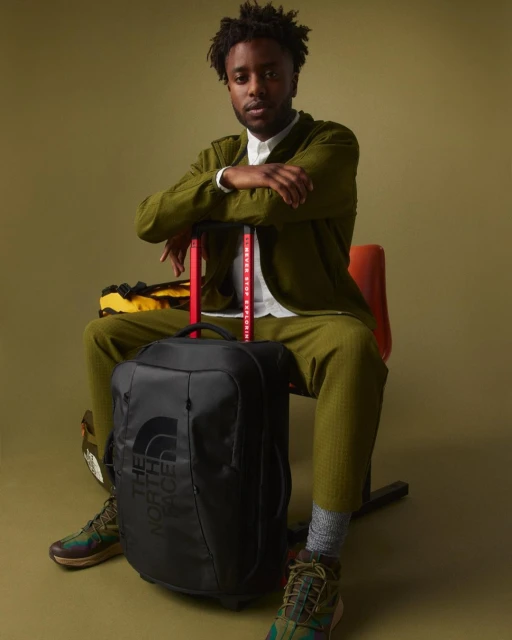 A seated adult Black man in a green suit with a black travel bag and The North Face branded gear, against a neutral green backdrop.