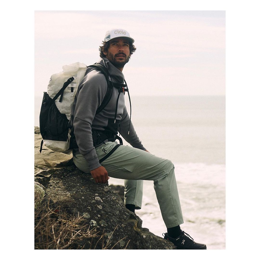 A man wearing a backpack and hat sits on a rock formation looking out to the ocean. The man is wearing outdoorsy active wear.