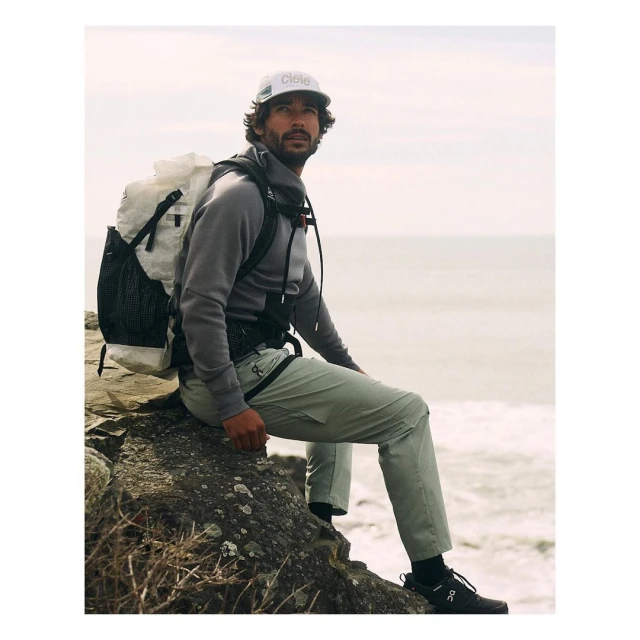 A man wearing a backpack and hat sits on a rock formation looking out to the ocean. The man is wearing outdoorsy active wear.