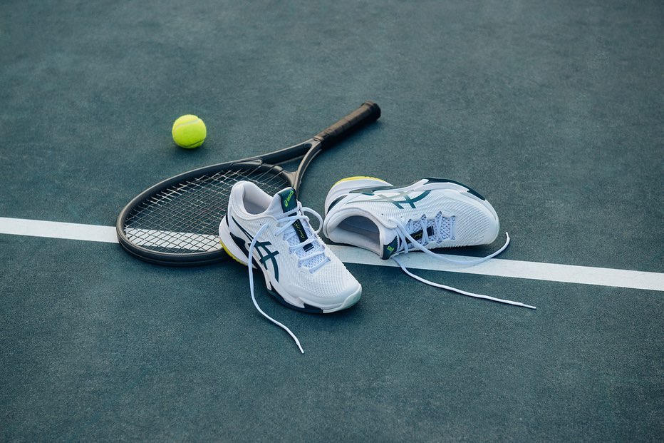 Overhead shot of tennis shoes, a racket, and a tennis ball on a blue tennis court with white lines. Athletic footwear close-up.