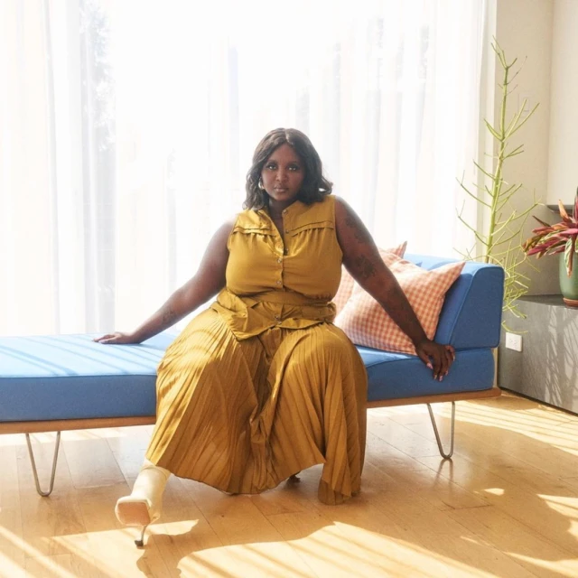 Woman in mustard sleeveless top and pleated skirt poses on blue mid-century modern bench with a light background and hardwood floors.