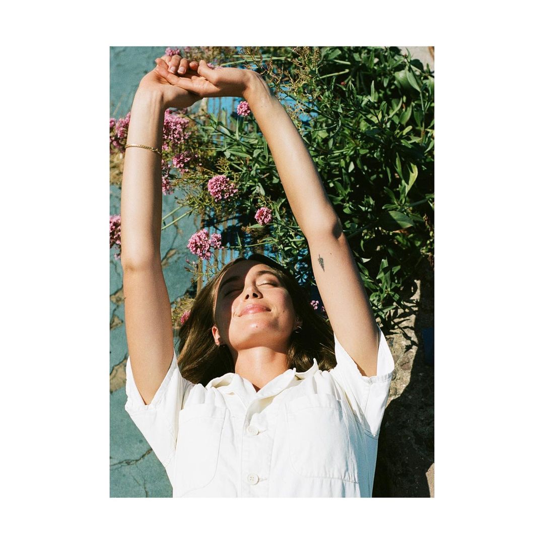 Woman in a white jumpsuit lies on blue concrete surrounded by green plants with pink flowers, arms raised, lit by warm sunlight.