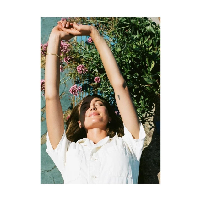 Woman in a white jumpsuit lies on blue concrete surrounded by green plants with pink flowers, arms raised, lit by warm sunlight.