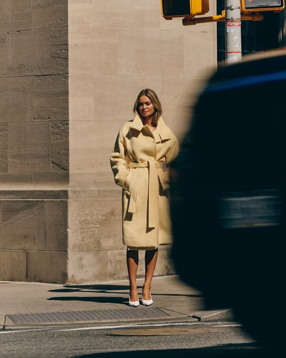 A woman in a yellow belted coat poses on a city street with a blurred car. The image focuses on fashion and urban lifestyle.