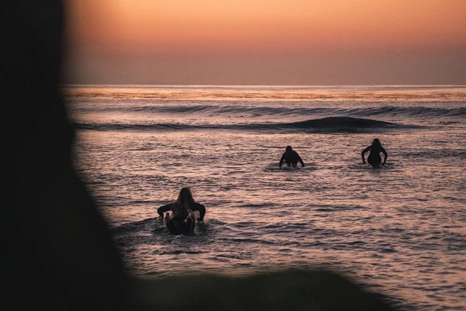 Three surfers wading in the ocean at dusk with a pink and orange sky, enjoying a day of watersports under the setting sun.