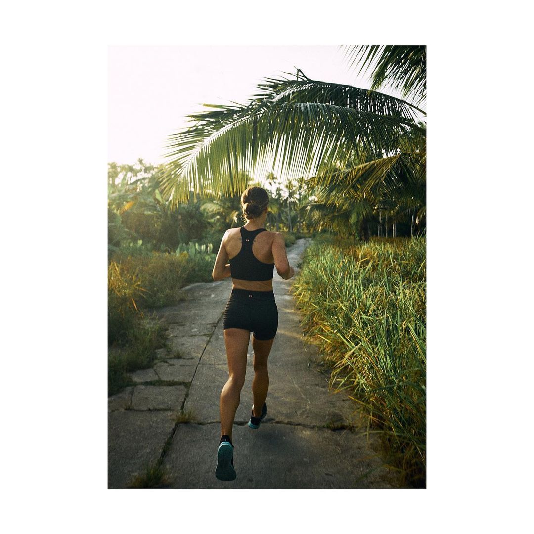 Athletic woman runs on a stone path lined with tropical plants under a sunny sky, showcasing fitness and outdoor lifestyle.