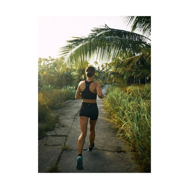 Athletic woman runs on a stone path lined with tropical plants under a sunny sky, showcasing fitness and outdoor lifestyle.