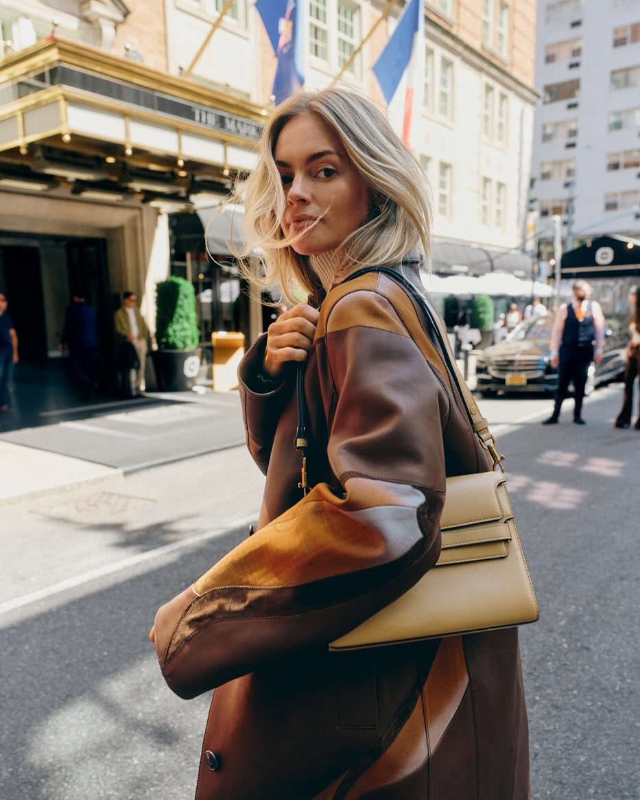 A stylish woman in a patterned coat walks down a city street with buildings and a hotel visible in the background, carrying a handbag.