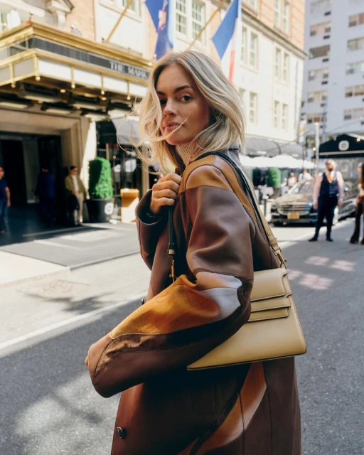 A stylish woman in a patterned coat walks down a city street with buildings and a hotel visible in the background, carrying a handbag.