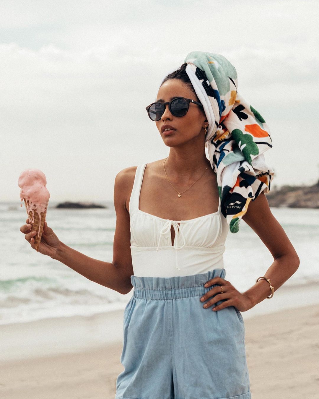 A woman on a beach holds a melting ice cream cone; she wears a colorful towel on her head and looks to the side, summery and stylish.