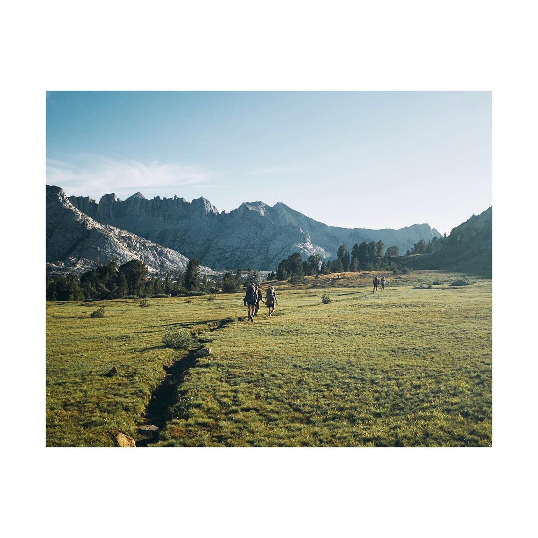 Hikers traverse a lush green meadow toward jagged mountains under a clear sky, capturing a nature and adventure theme in a scenic landscape.