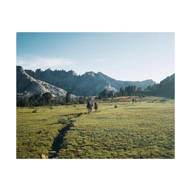 Hikers traverse a lush green meadow toward jagged mountains under a clear sky, capturing a nature and adventure theme in a scenic landscape.