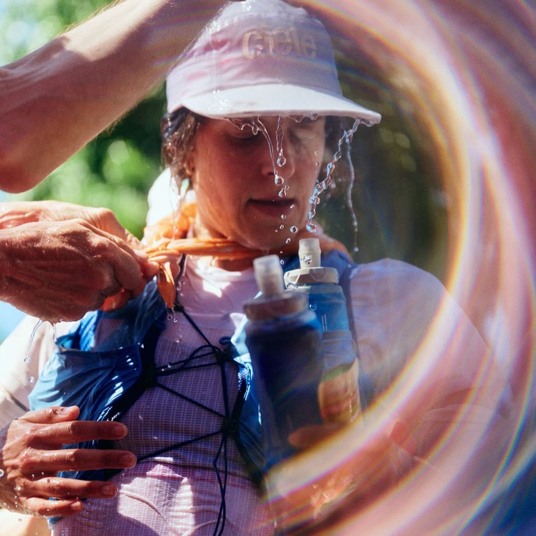 Adult woman in athletic gear gets water poured over her head by another person, likely during an outdoor sporting event, wearing a cap.