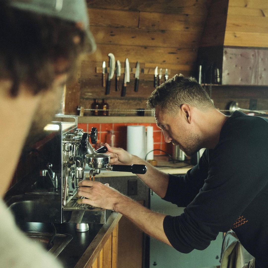A man operates a chrome espresso machine in a rustic indoor setting, another man observes, with knives on a wood background.
