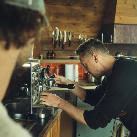 A man operates a chrome espresso machine in a rustic indoor setting, another man observes, with knives on a wood background.