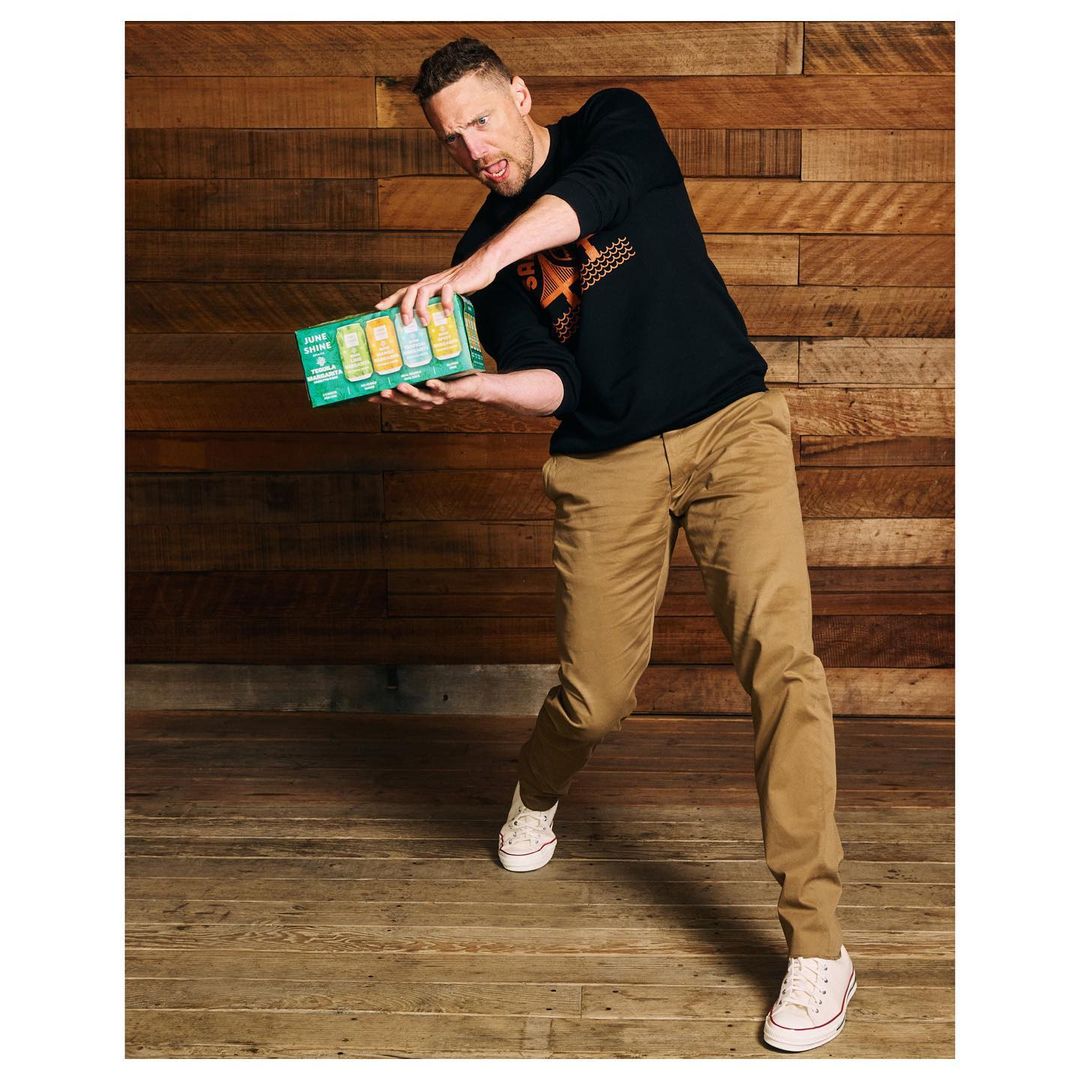 Adult male holds a pack of beverages against a wood background, wearing casual clothing and white shoes in an indoor setting.