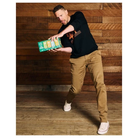 Adult male holds a pack of beverages against a wood background, wearing casual clothing and white shoes in an indoor setting.