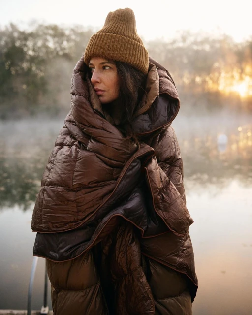Woman wrapped in a brown puffer blanket wearing a beanie stands outside near water with trees in the misty background.
