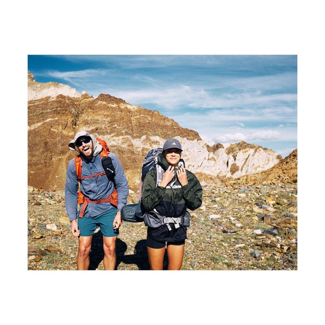 Two backpackers, a man and a woman, stand smiling in a rocky, mountainous landscape under a partly cloudy sky, enjoying their hike.
