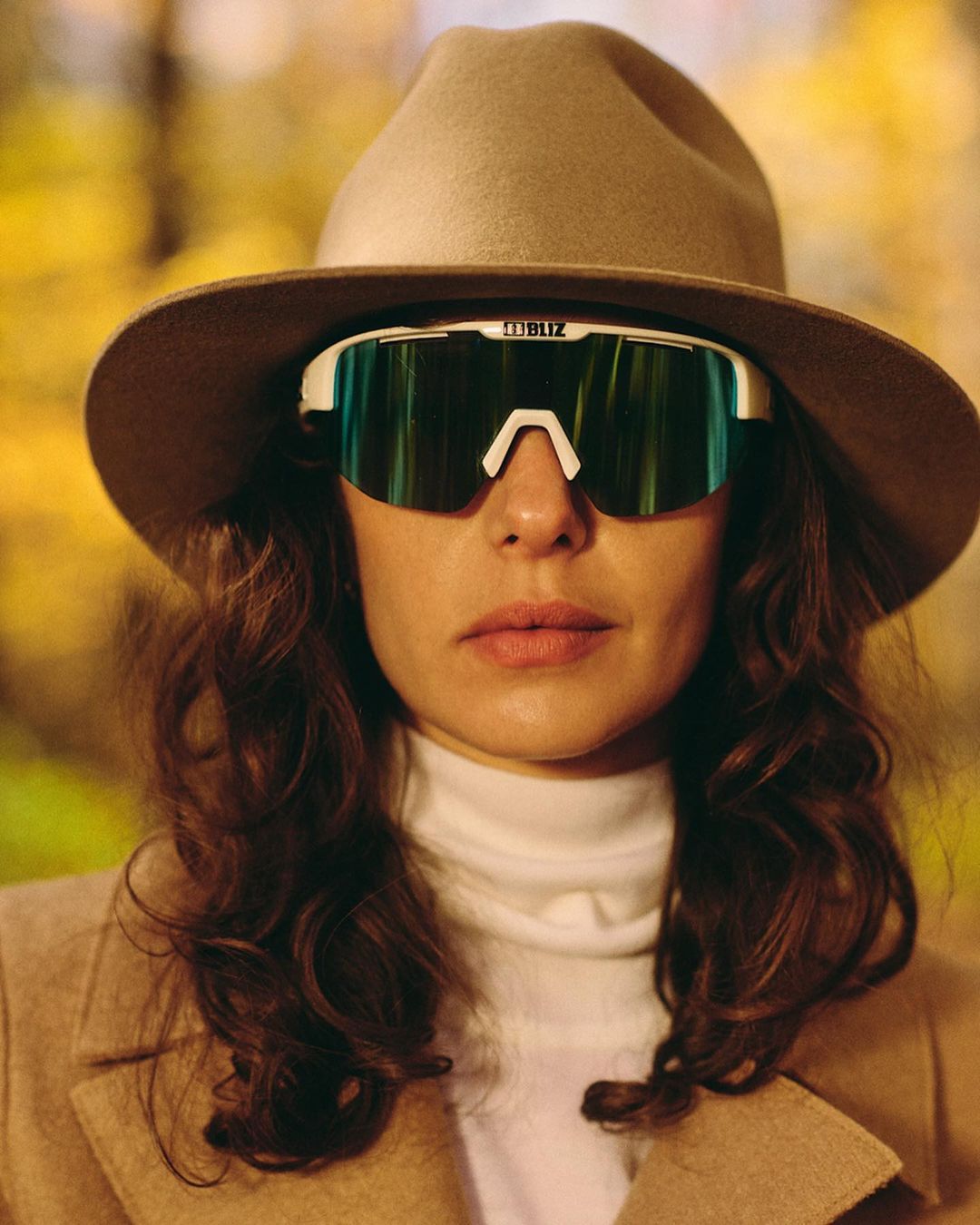 Portrait of an adult woman wearing sunglasses and a hat, looking directly at the camera with an outdoor, blurred background.