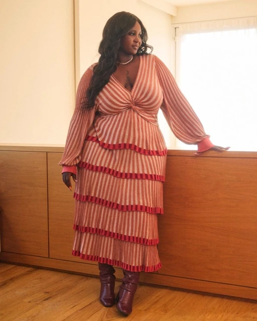 An African American woman with long dark hair wearing a striped dress and boots posing indoors with a wood cabinet in the background.