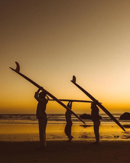 Three silhouetted people on a beach carry surfboards on their heads at sunset, with golden light reflecting on the wet sand.