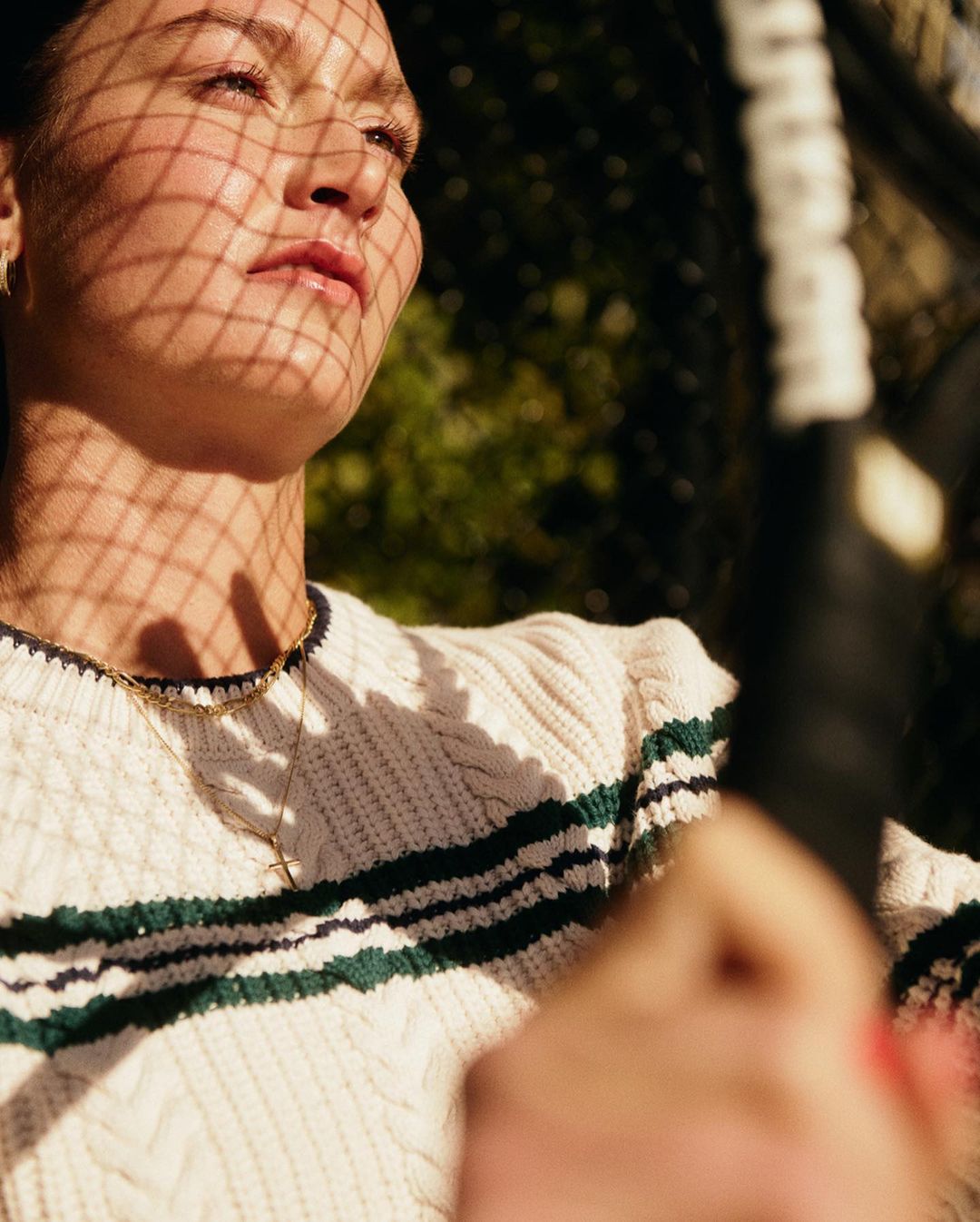 Portrait of a woman in a cream cable knit sweater holding a tennis racket, her face patterned with shadow from a net.