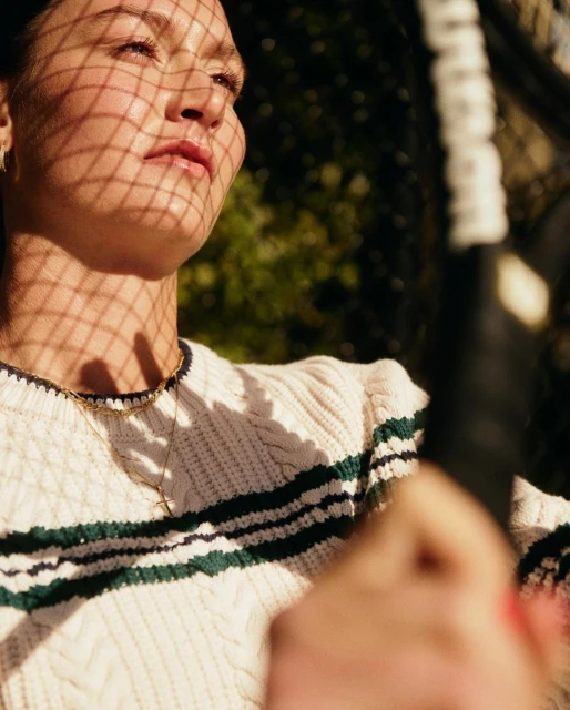 Portrait of a woman in a cream cable knit sweater holding a tennis racket, her face patterned with shadow from a net.