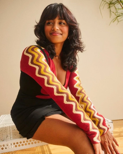 Portrait of a woman with dark hair in a black dress and patterned sweater, sitting and smiling in an indoor setting with natural light.