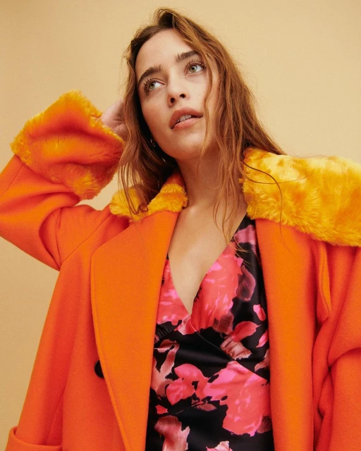 Woman in an orange coat with faux fur collar and cuffs, wearing a floral dress against a tan backdrop in a studio shot.