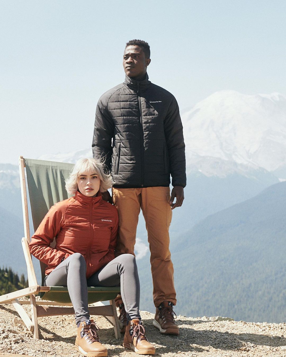 A man and woman in outdoor apparel are posing on a hillside with a mountain backdrop. They are wearing matching boots and jackets.