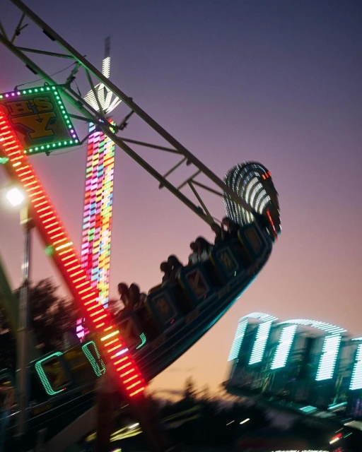 An amusement park ride spins with lights blurring at dusk; a colorful tower rises in the background against a gradient sky.