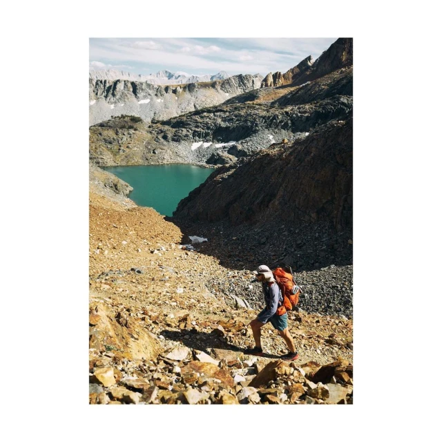A person with a backpack hikes up a rocky mountain path towards a lake surrounded by rocky peaks under a bright, partly cloudy sky.