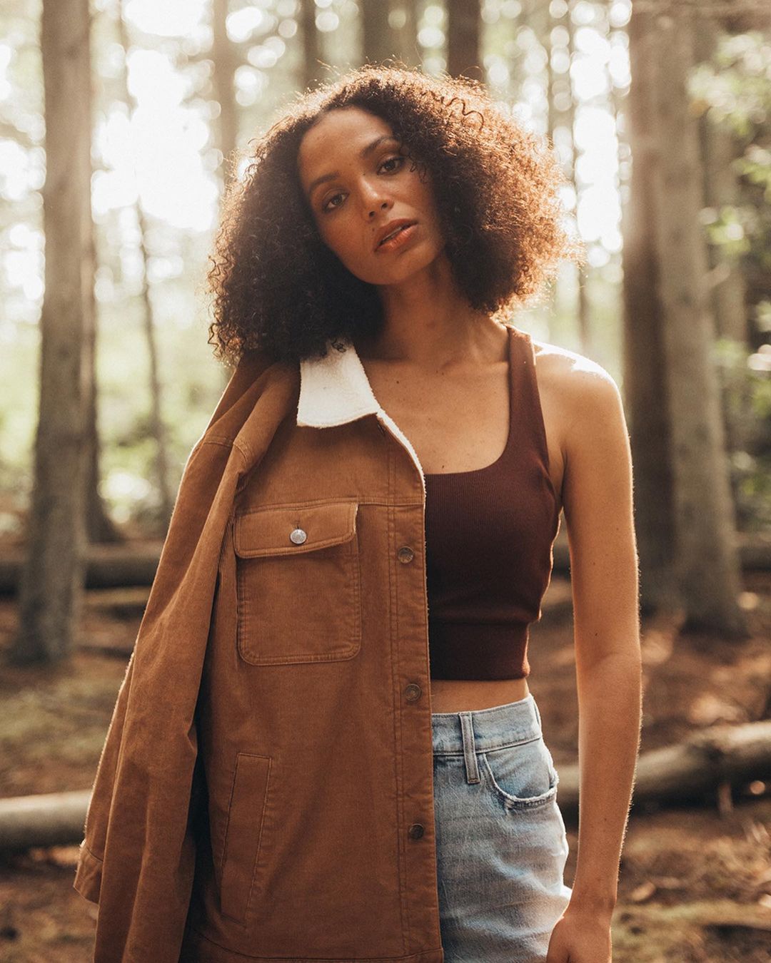 A woman with curly hair poses outdoors wearing a brown jacket and jeans, surrounded by a forest setting with soft, diffused lighting.