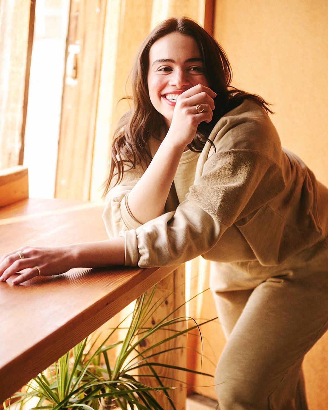 Portrait of a smiling woman in a light-brown sweatsuit leaning against a wooden counter, with a houseplant in the background.