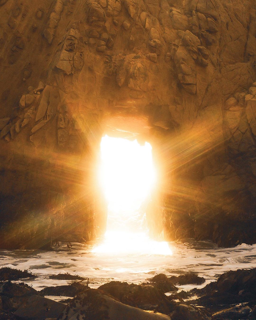 Cave opening overlooking the sea with bright light shining through the entrance. Rock formations surround the illuminated view to the ocean.