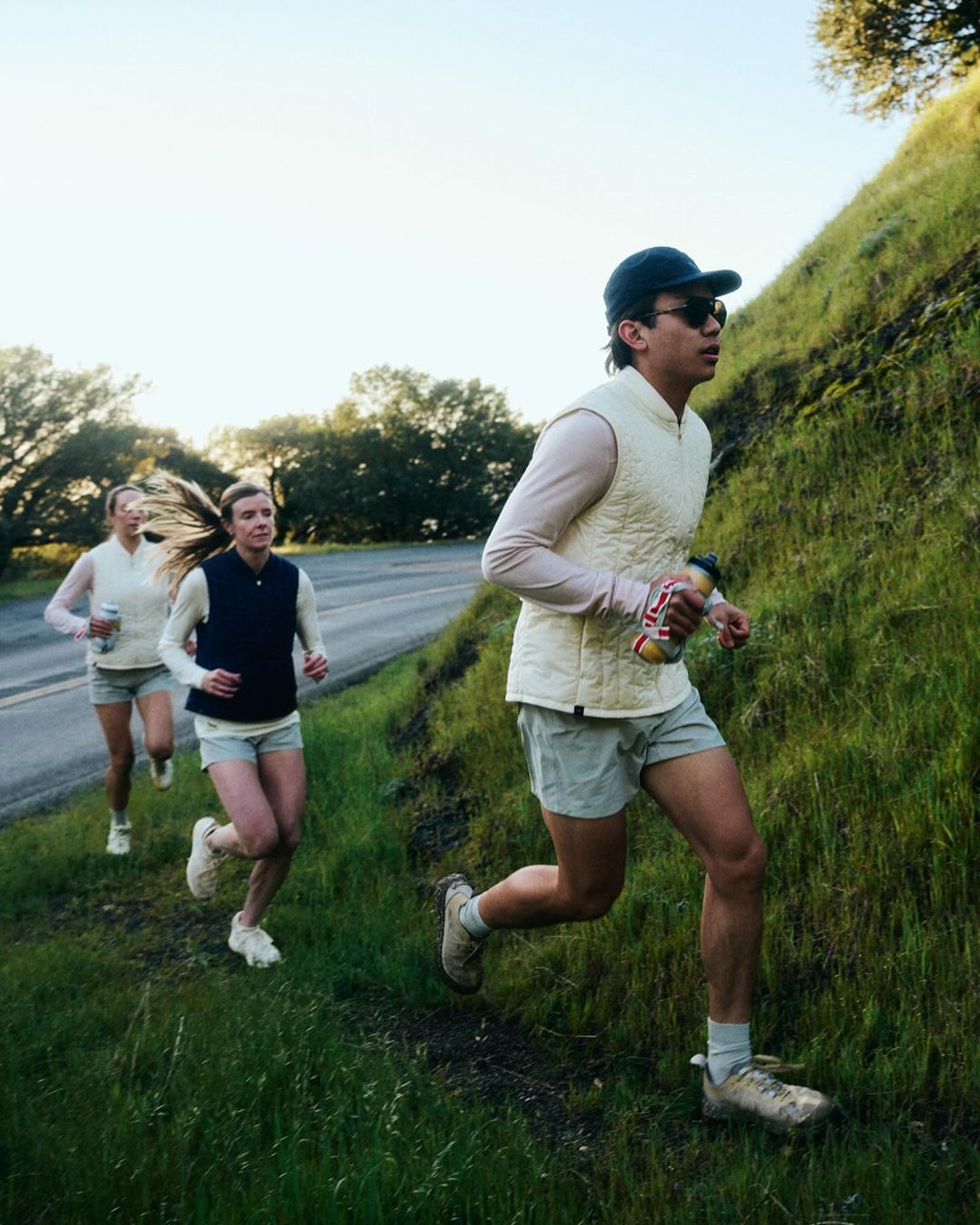 Three adult women and one adult male trail running on a grassy hillside next to an asphalt road wearing athletic apparel and carrying water bottles.