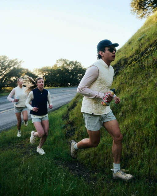Three adult women and one adult male trail running on a grassy hillside next to an asphalt road wearing athletic apparel and carrying water bottles.