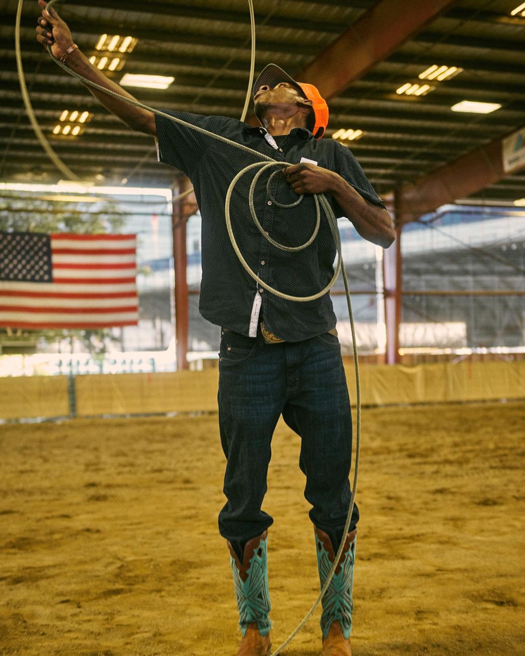 A man in jeans and cowboy boots throws a lasso high in the air during a rodeo performance indoors, with the American flag in the background.